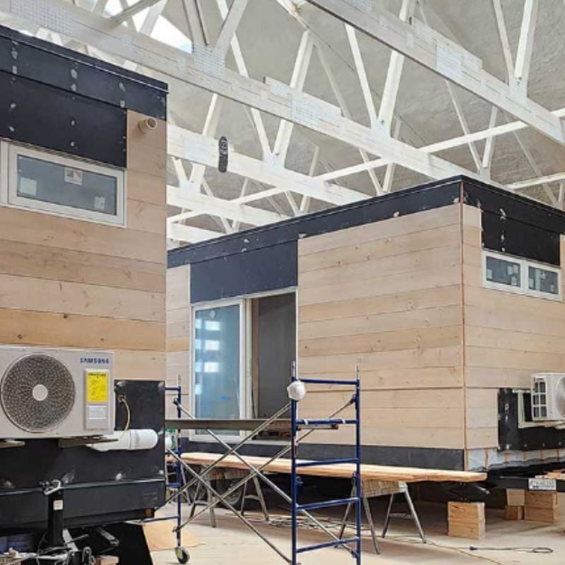 Two WheelPad accessible modular homes under construction inside a manufacturing facility, showcasing modern wood-and-black exteriors, integrated accessibility features, and an efficient off-site building process designed to create high-quality, accessible living spaces.