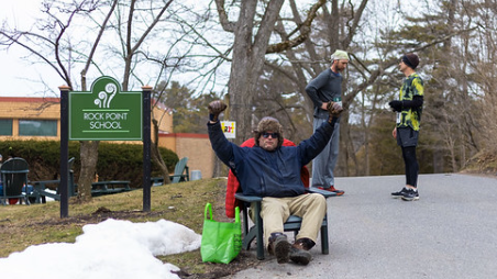 A person in a winter jacket and big hat sit by a Rock Point School sign on campus waiting for finishers of the Maple Run 5k Race
